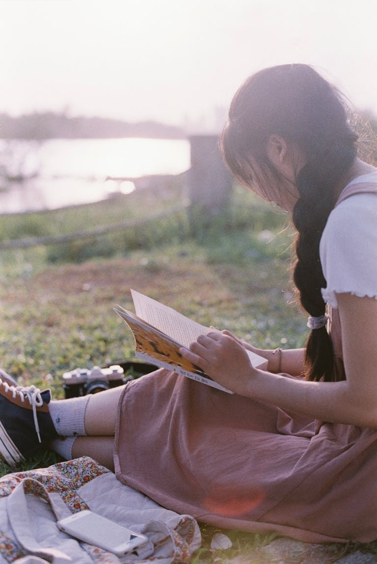 Girl reading a book by a riverside at sunset, creating a serene summer scene.